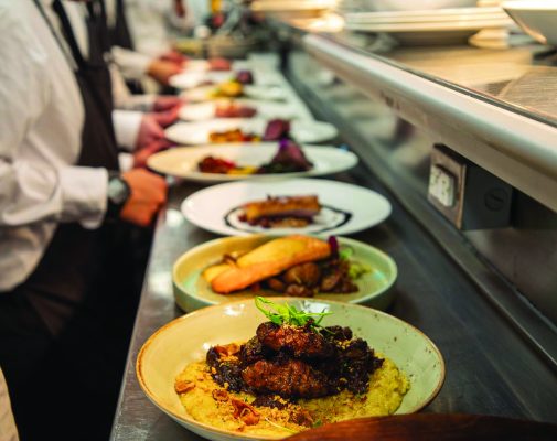 A row of plated gourmet dishes sits on a kitchen counter at Terrapin Restaurant, ready to be served by staff in white shirts and aprons. The focus is on the front plate, which features a meat dish on a bed of grains.