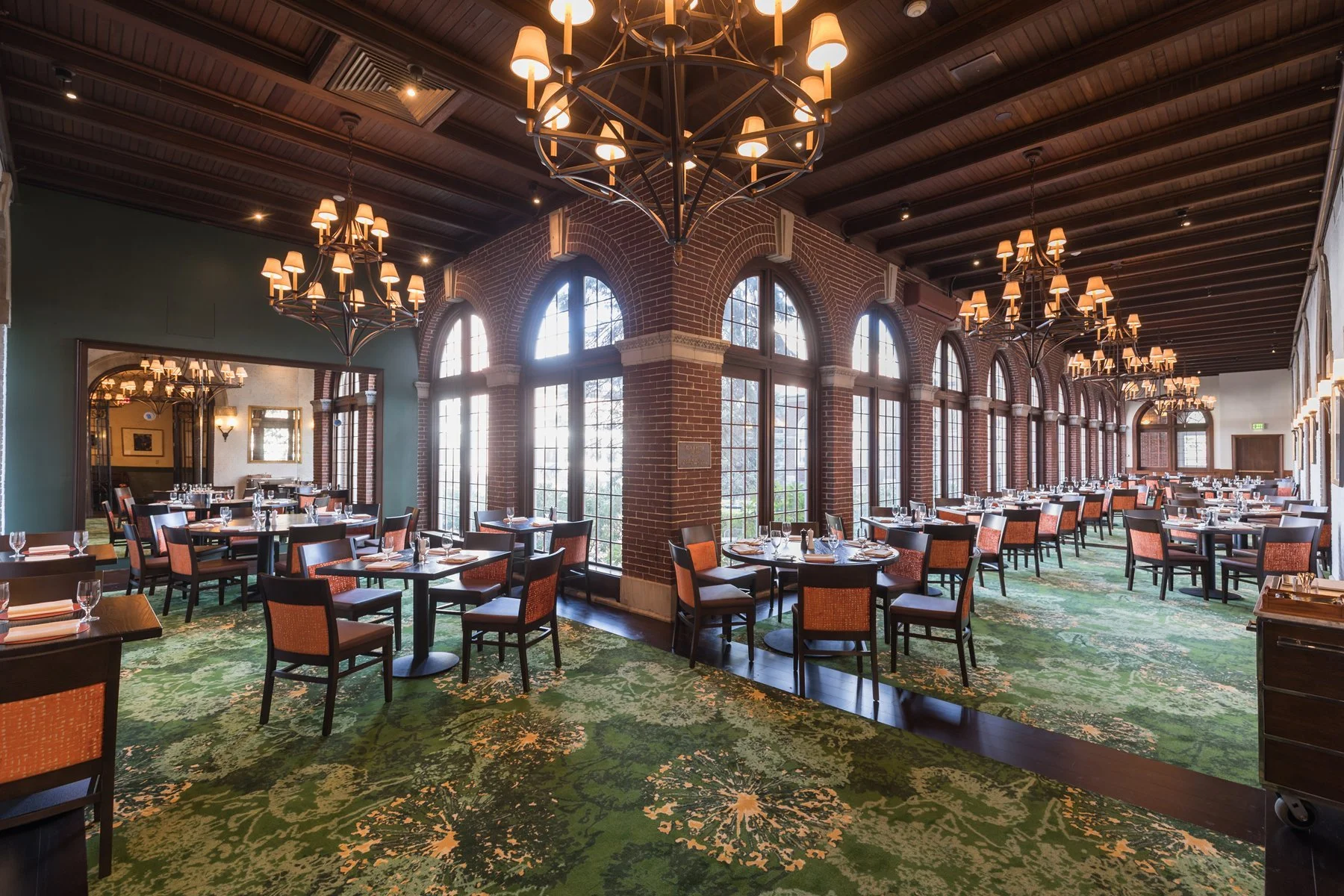 Spacious American Bounty Restaurant with arched windows, brick walls, green patterned carpet, wooden ceiling, elegant chandeliers, and neatly set tables with orange chairs. Natural light streams in, creating a warm, inviting atmosphere.