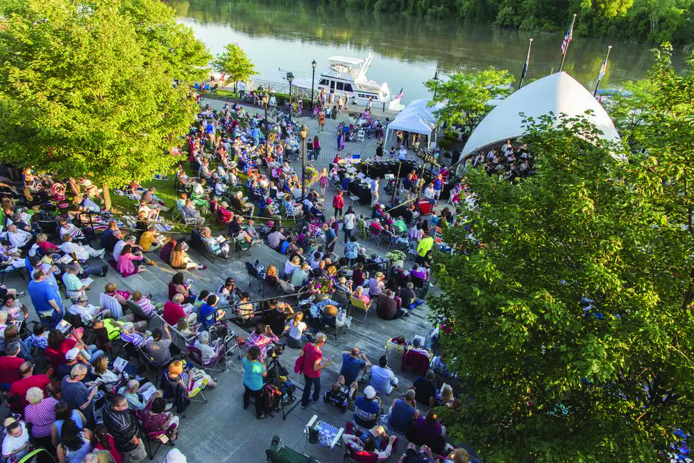 A large crowd sits outdoors near a river, facing a stage with a white tent. People are seated on steps and grass, surrounded by green trees, enjoying a live performance. Boats are docked in the background, while visitors savor unique drinks from the Adirondack Craft Beverage Trail for a truly local experience.