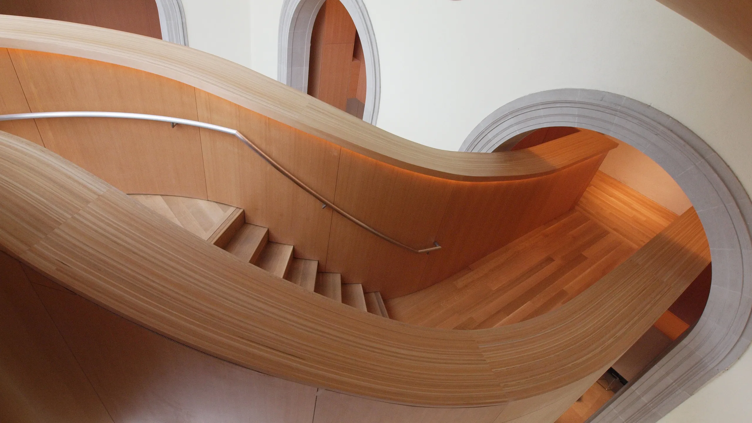Curving wooden staircase with a metal handrail, light wood steps, and arched doorways in a modern, minimalist interior space reminiscent of the Art Gallery of Ontario.