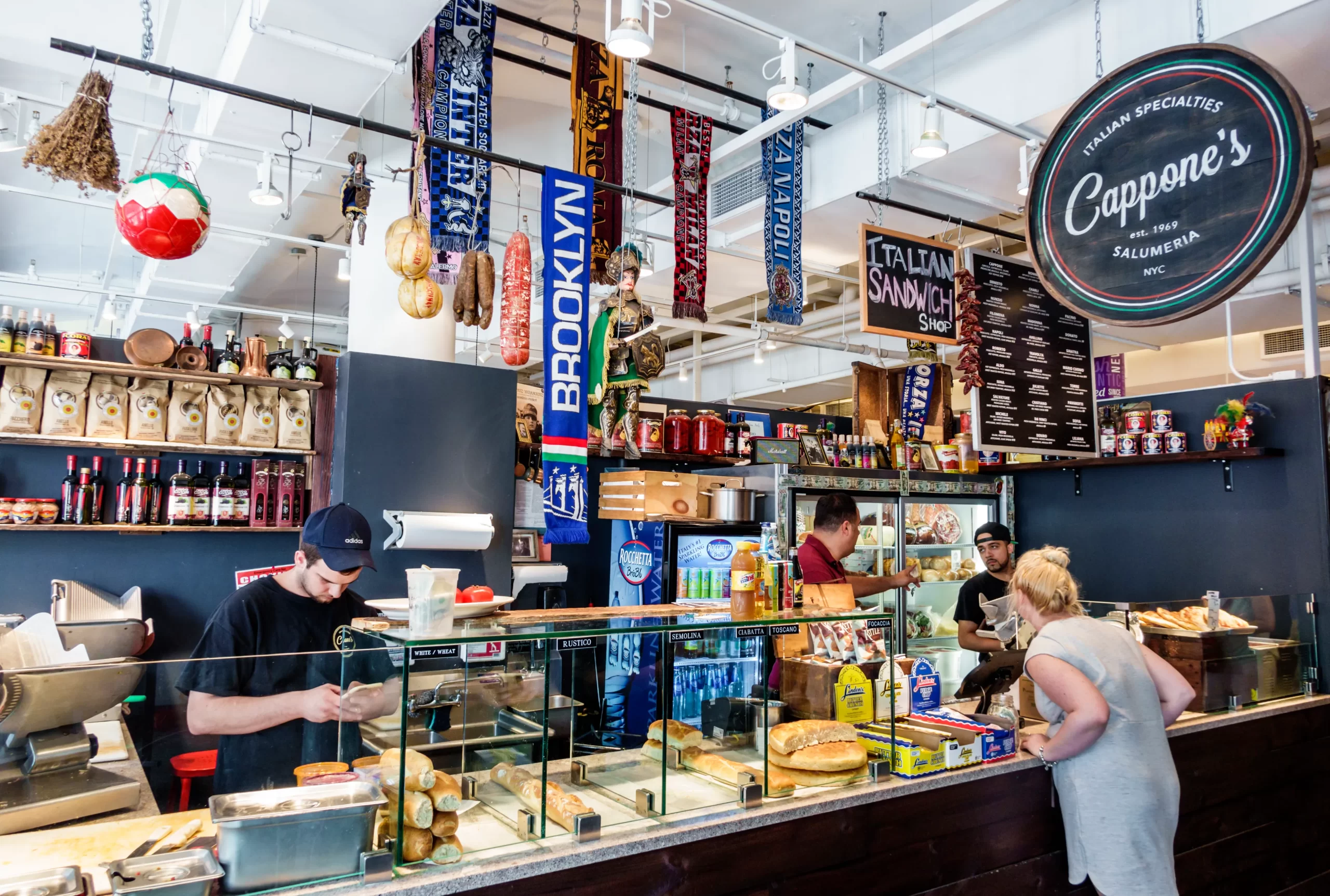 A busy deli counter at Chelsea Market with staff preparing sandwiches, shelves stocked with Italian goods, and a customer ordering. Signs, sausages, and food adorn the bright, bustling space.