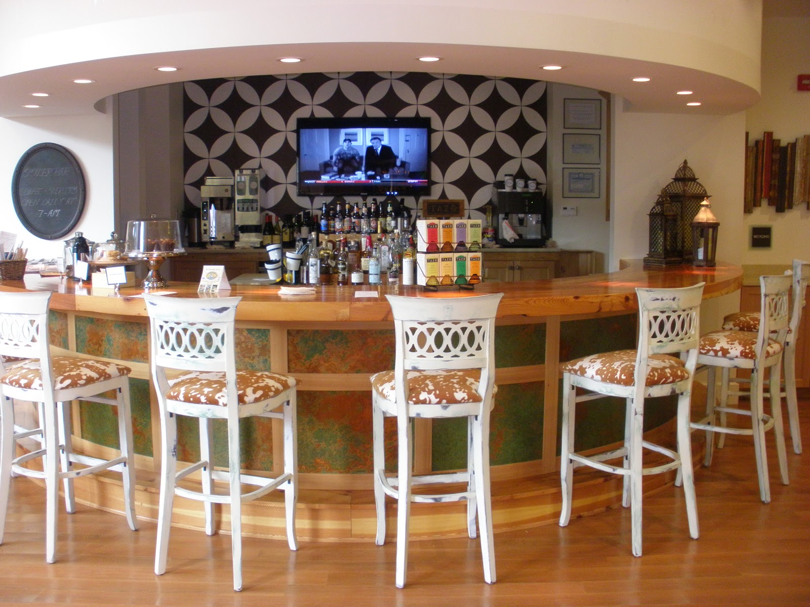 A curved bar with five white barstools, various bottles and glasses, a patterned wall, a TV screen, and warm lighting create a cozy ambiance at Hotel Skyler Syracuse.