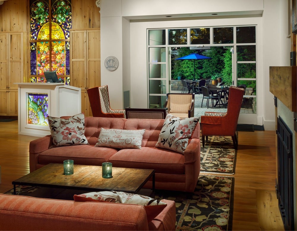 Cozy living room with red sofas, floral pillows, and a stained glass window evokes the inviting atmosphere of Hotel Skyler Syracuse. Glass doors open to a patio with outdoor seating and a blue umbrella, surrounded by greenery.