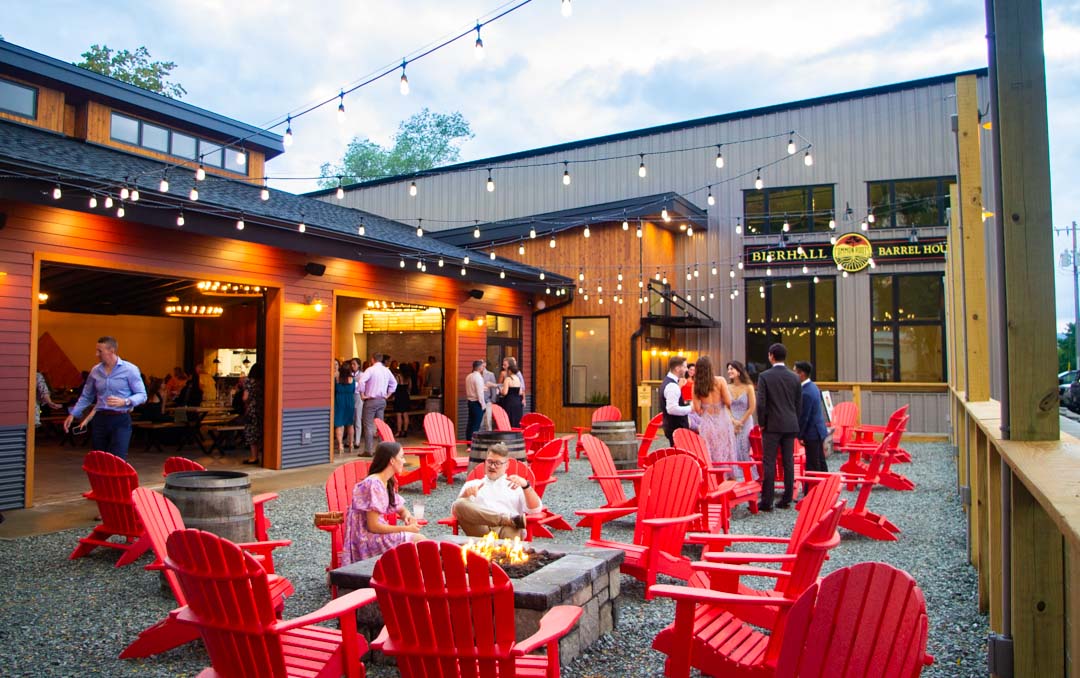 People gather in an outdoor patio area with red Adirondack chairs, a fire pit, and string lights, beside a modern building that reads Barrel House. Whether talking or relaxing by the fire, visitors enjoy the welcoming atmosphere—a perfect stop along the Adirondack Craft Beverage Trail.