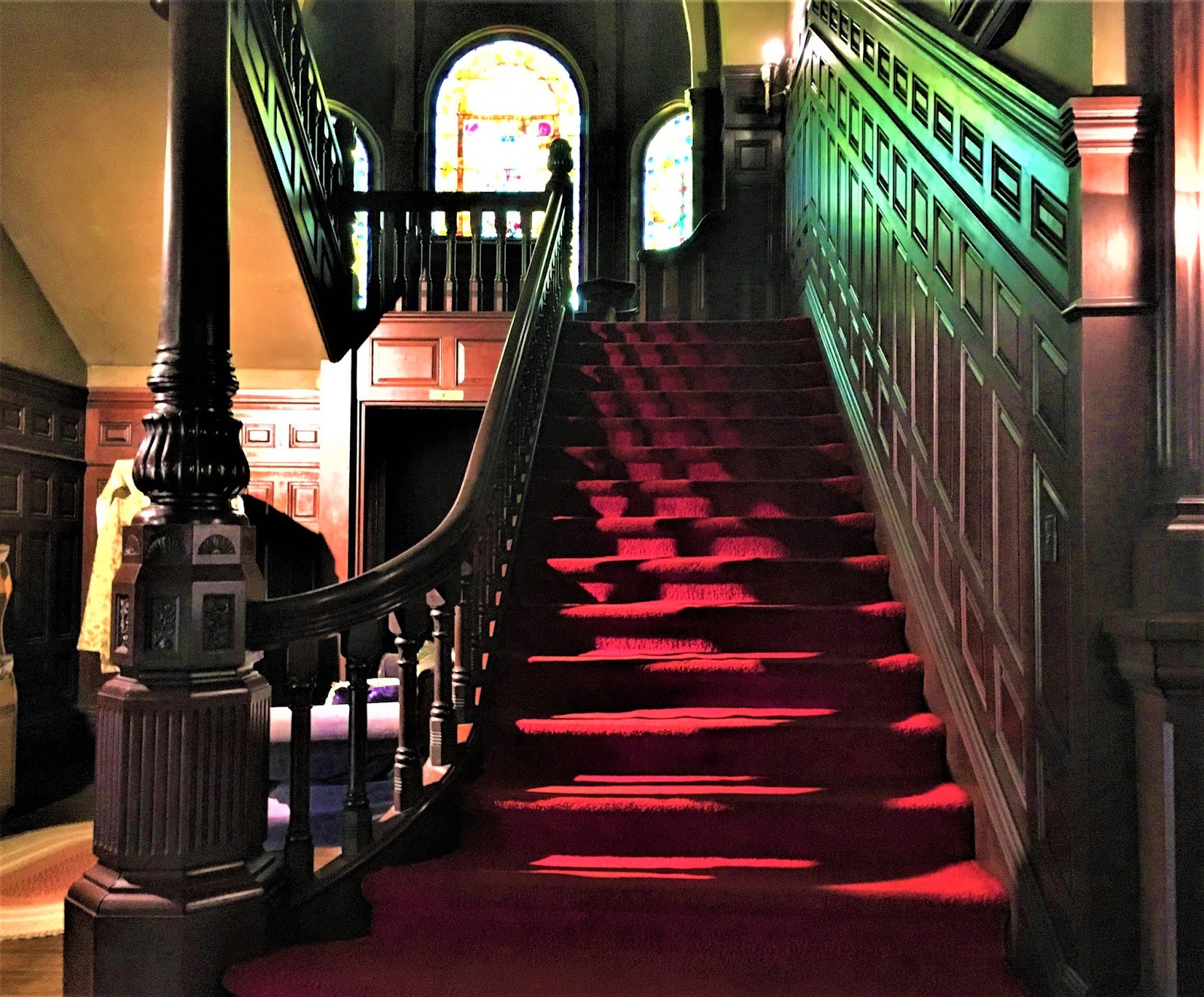 A grand wooden staircase with a red carpet runner leads up to stained glass windows, surrounded by ornate wood paneling and dim, ambient lighting in the historic Wilson Castle.