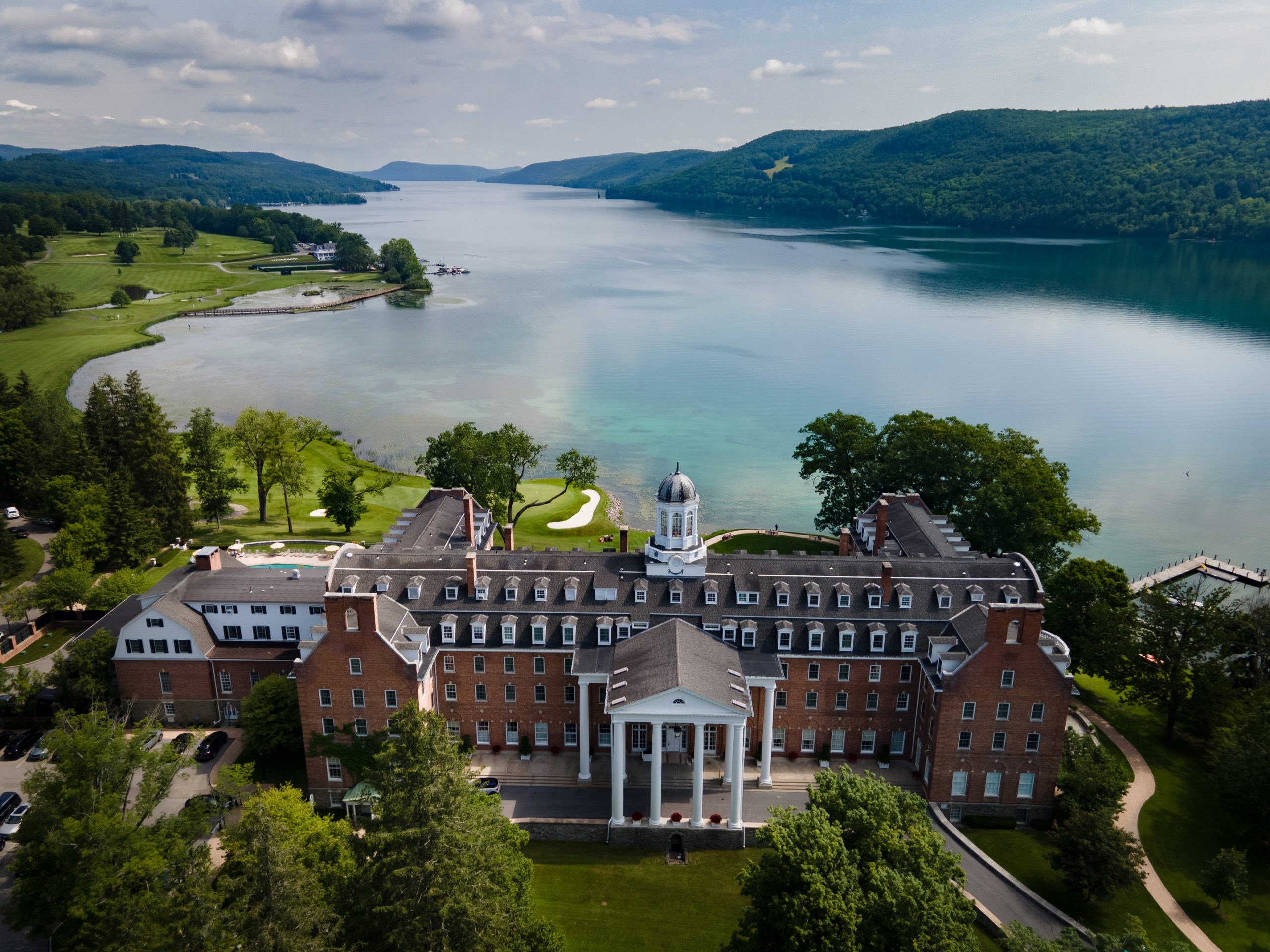 Aerial view of The Otesaga Resort Hotel, a large brick building with a white dome, set by a lake with hills and trees in the background on a partly cloudy day.