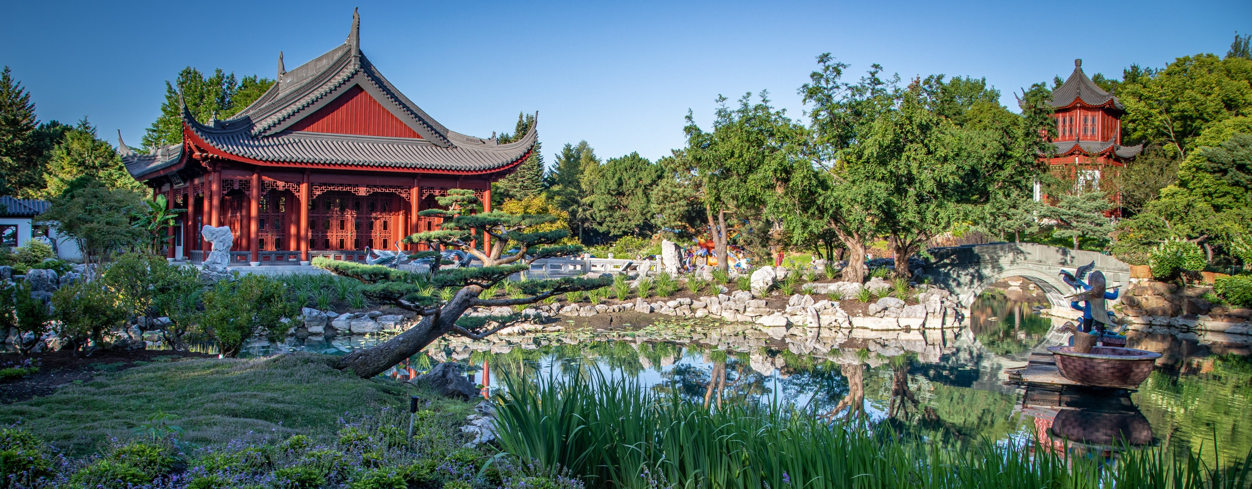 A traditional red pagoda stands by a serene pond surrounded by lush greenery, rocks, and a small arched bridge under a clear blue sky, reminiscent of the tranquil landscapes found in the Jardin Botanique De Montréal.