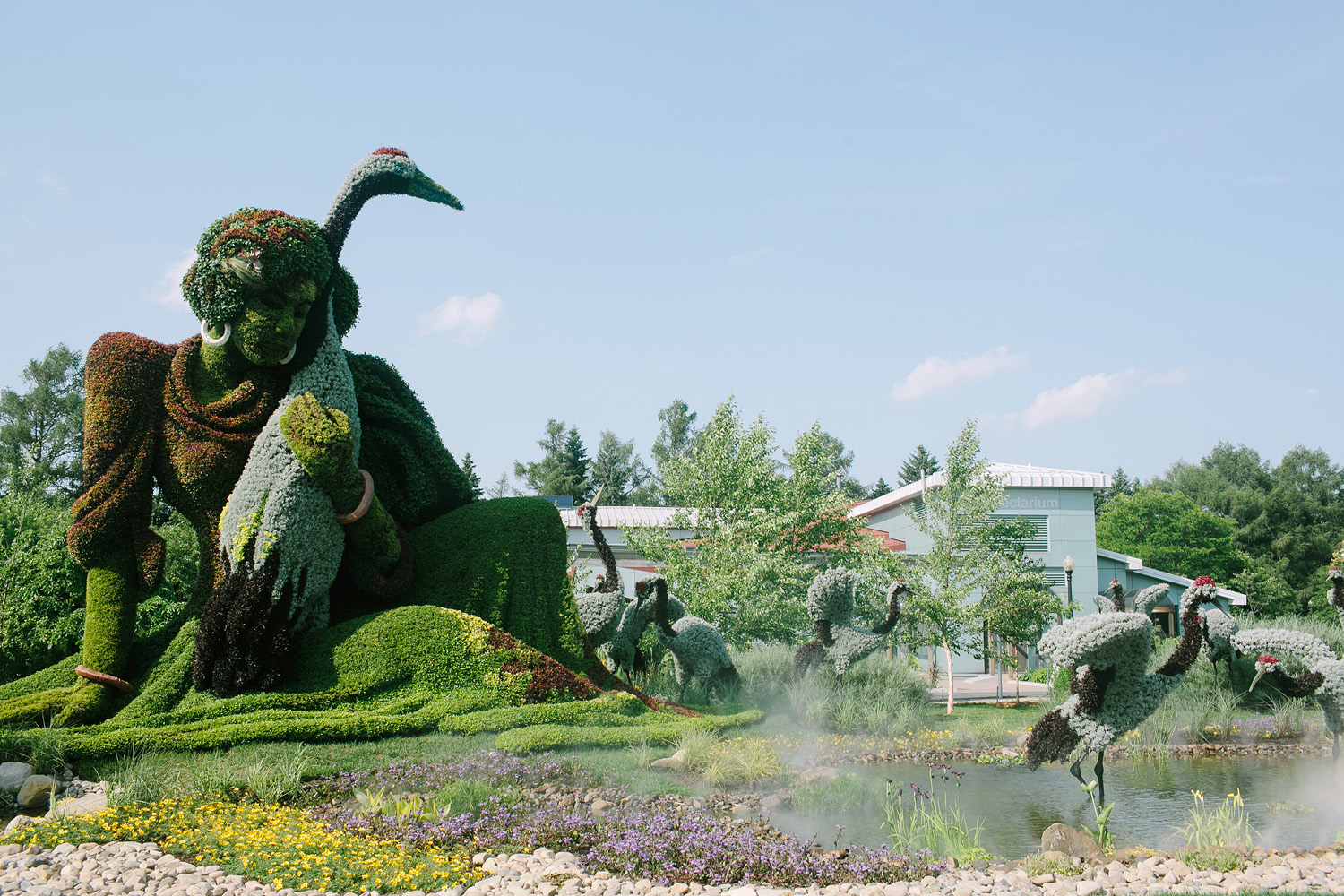 A large topiary sculpture of a person with birds, surrounded by greenery, flowers, and a pond under a clear sky, with buildings and trees in the background—reminiscent of the enchanting displays found at Jardin Botanique De Montréal.