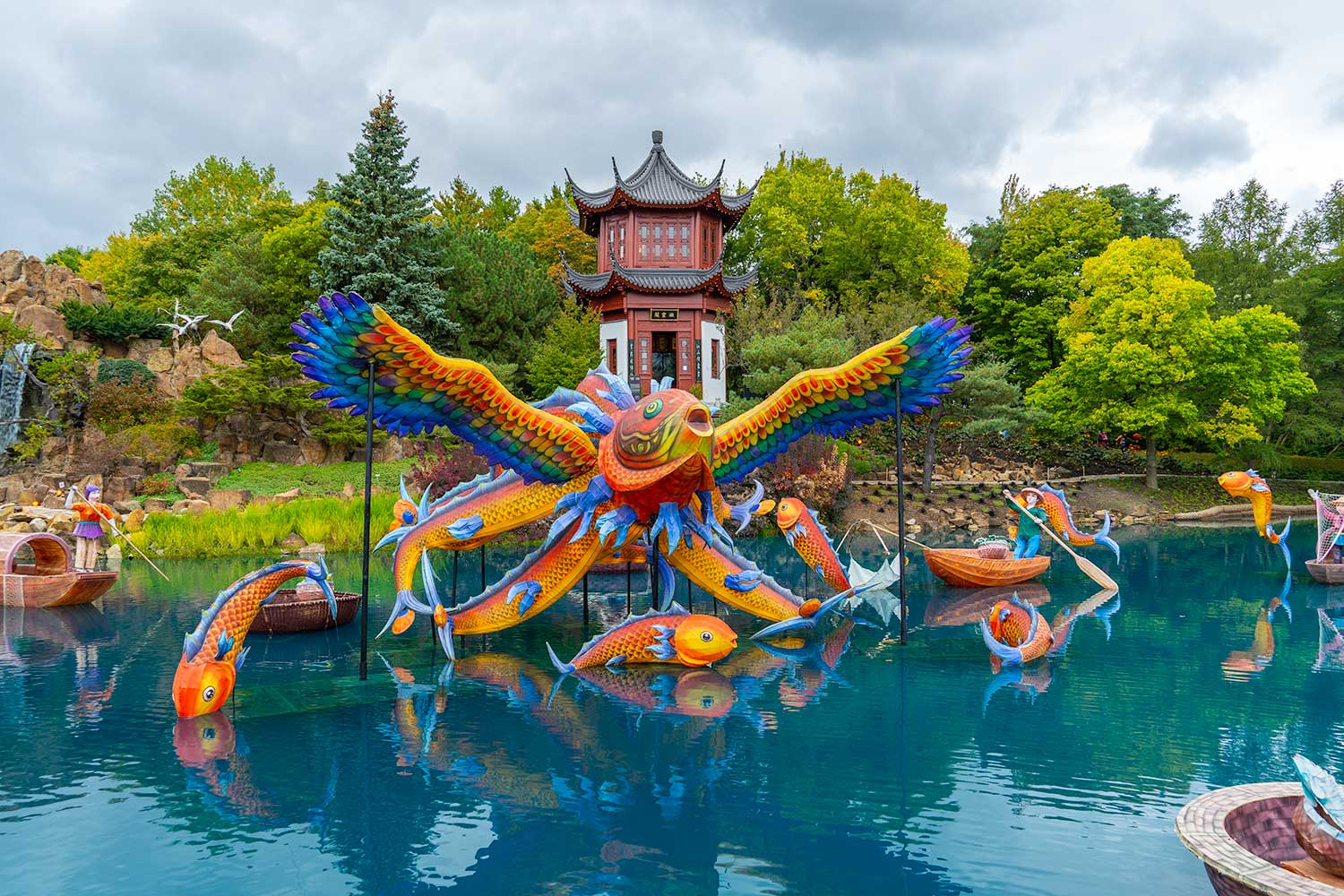 Colorful lantern sculptures of a phoenix, fish, and boats float on a pond near a traditional Chinese pavilion at the Jardin Botanique De Montréal, surrounded by lush greenery and rocks under a cloudy sky.