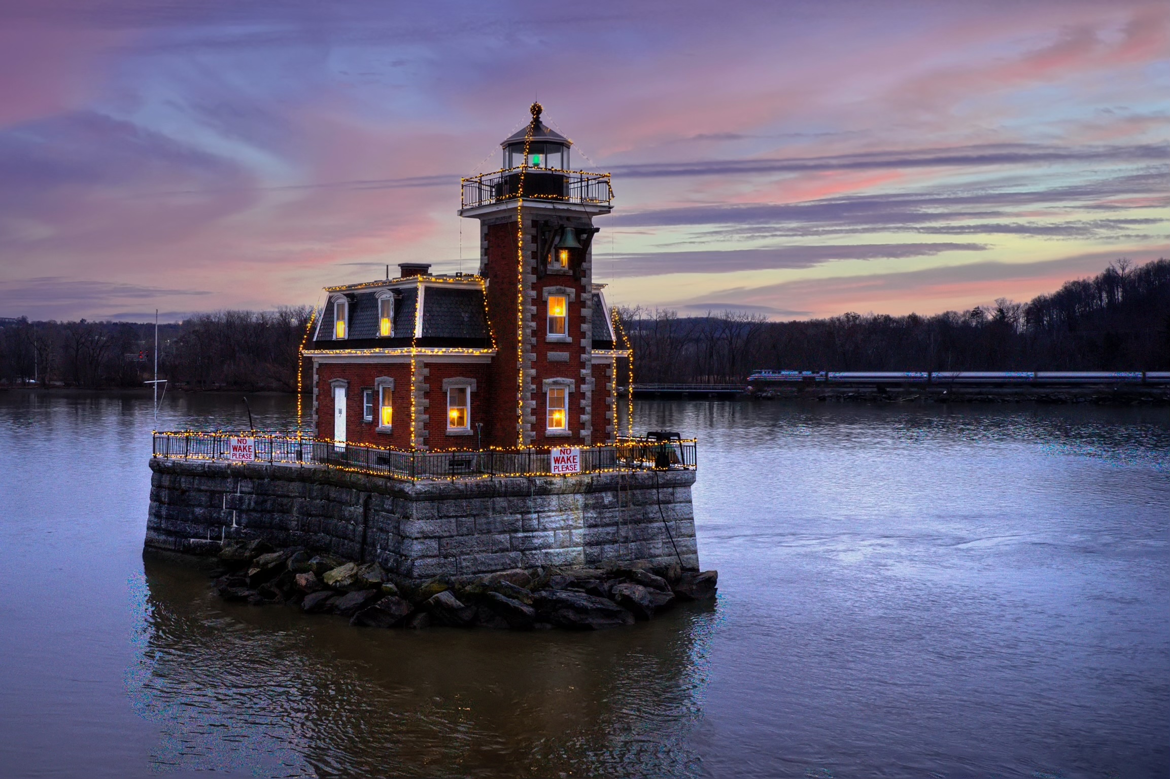 A small brick lighthouse, known as the Hudson-Athens Lighthouse, decorated with string lights sits on a stone base surrounded by water at sunset, with a colorful sky and trees in the background.