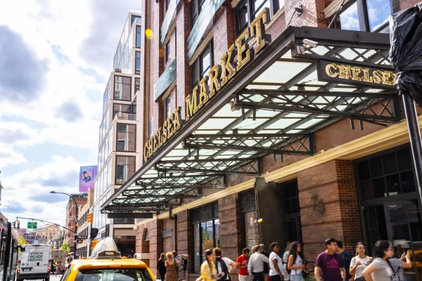 People walk under the large Chelsea Market sign in New York City on a busy street, with a yellow taxi and brick buildings visible under a partly cloudy sky—a scene that buzzes with energy reminiscent of the artistic gatherings at Caffè Lena.