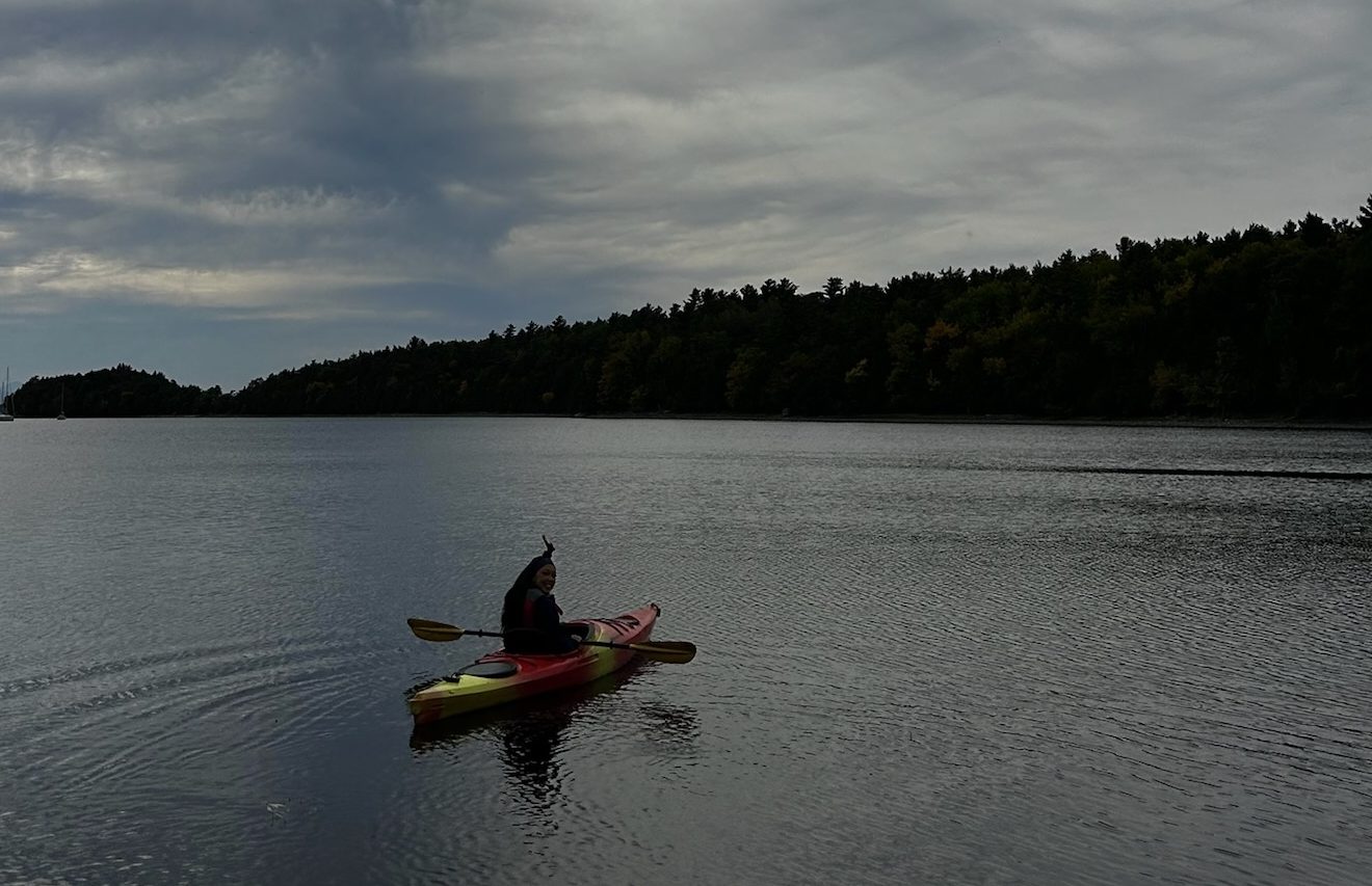 Miss New York explores Albany NY by Amtrak, then paddles a kayak on a calm lake under a dramatic sky with thick clouds, forested shoreline visible in the distance.