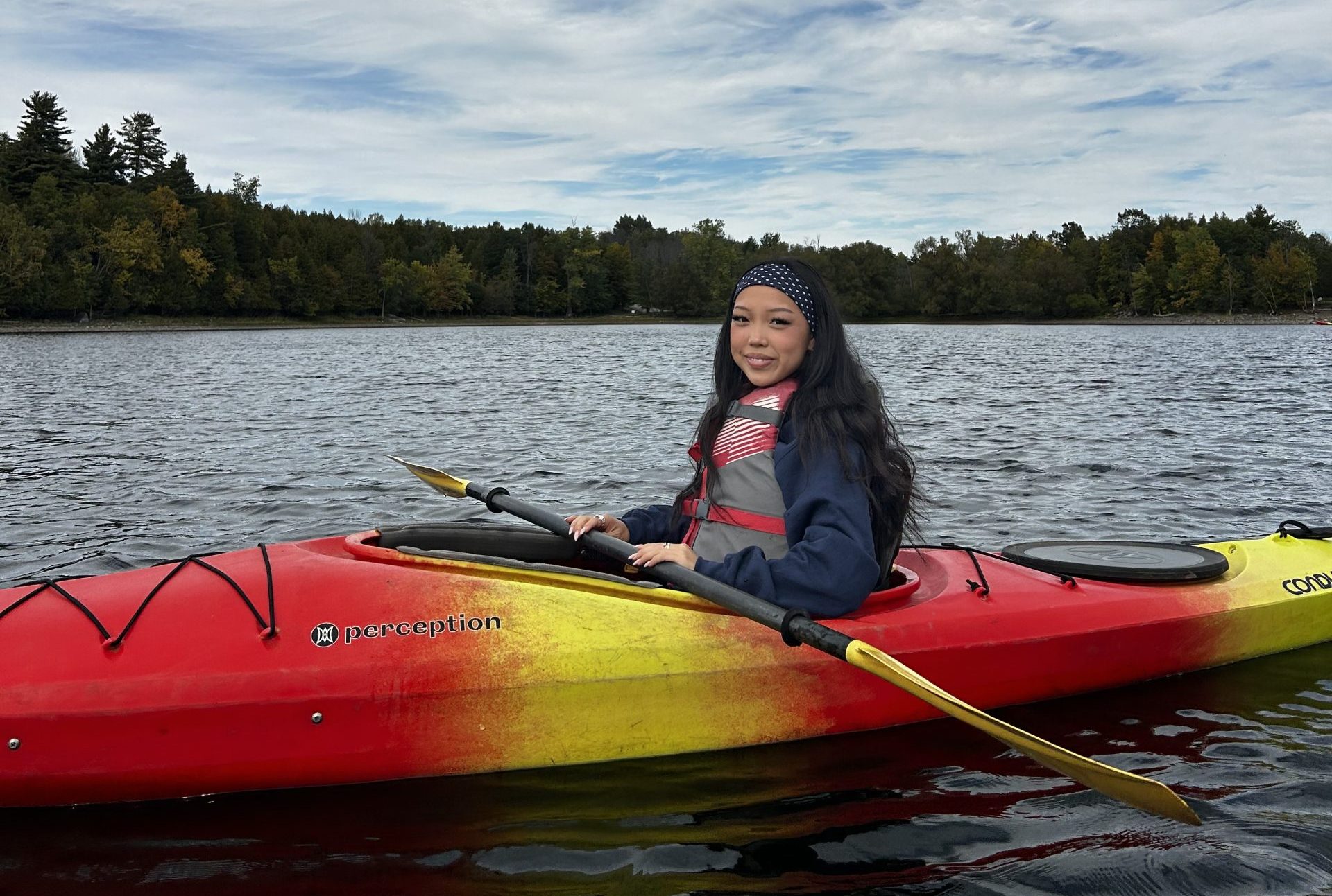 A person wearing a life jacket and knit hat sits in a red and yellow kayak on a lake, holding a paddle, as Miss New York explores Albany NY by Amtrak. Trees and a cloudy sky fill the background.