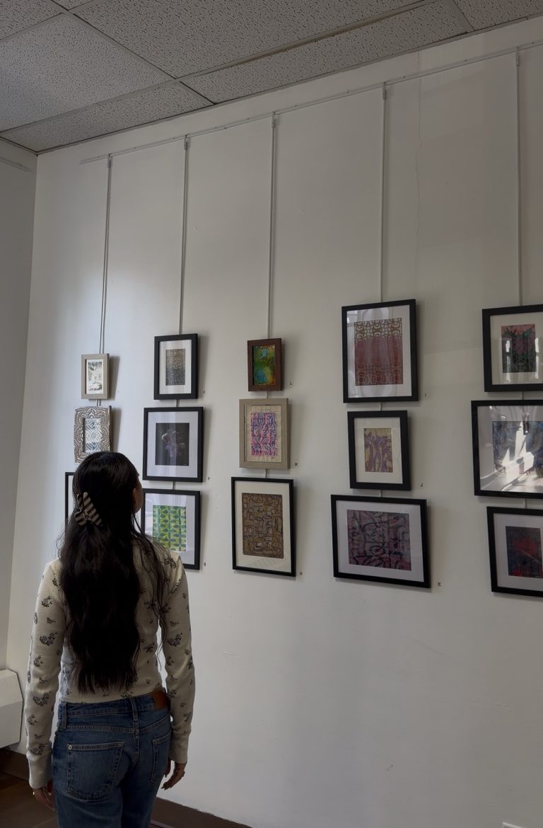 A person with long dark hair stands in front of a white wall displaying various framed artworks at an art gallery, reminiscent of scenes from Miss New York Explores Albany NY by Amtrak.