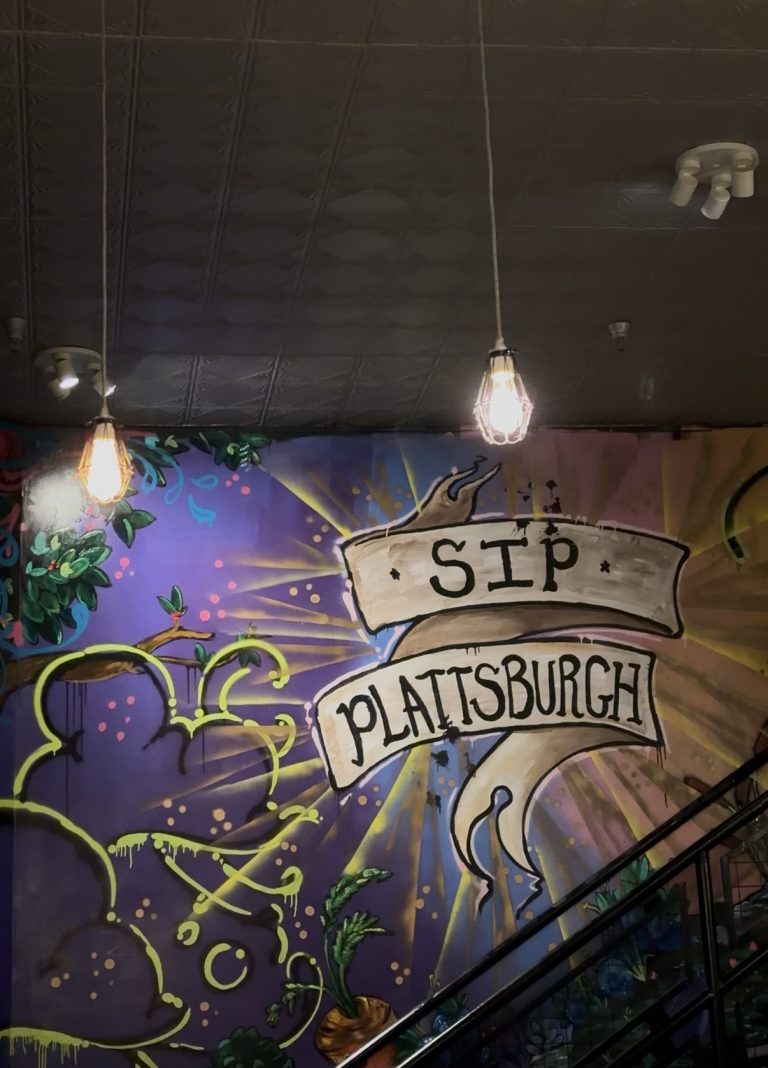 Colorful mural reading SIP PLATTSBURGH with sun rays, green vines, and hanging lightbulbs above a staircase and a purple background, reminiscent of Miss New York Explores Albany NY by Amtrak’s vibrant journeys.