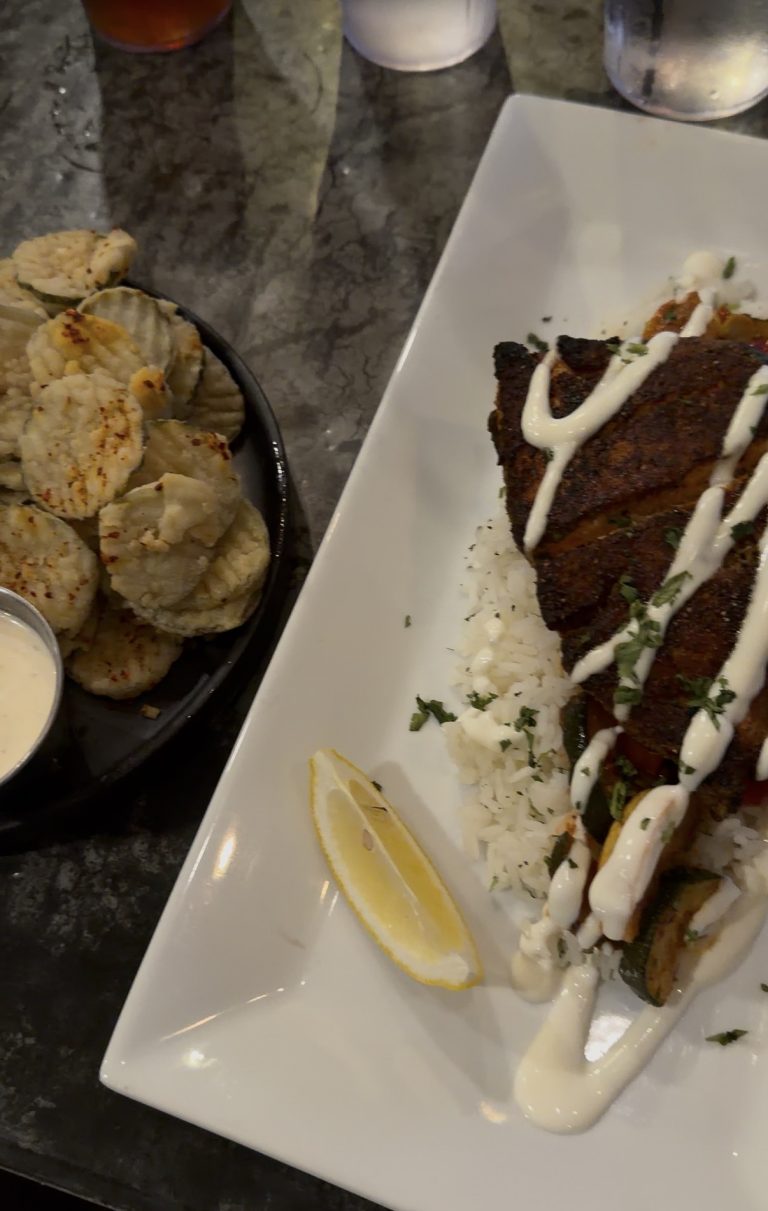 Miss New York Explores Albany NY by Amtrak: A plate with blackened fish on rice, drizzled with white sauce and herbs, plus fried pickles with dipping sauce—delicious flavors await your next rail adventure.
