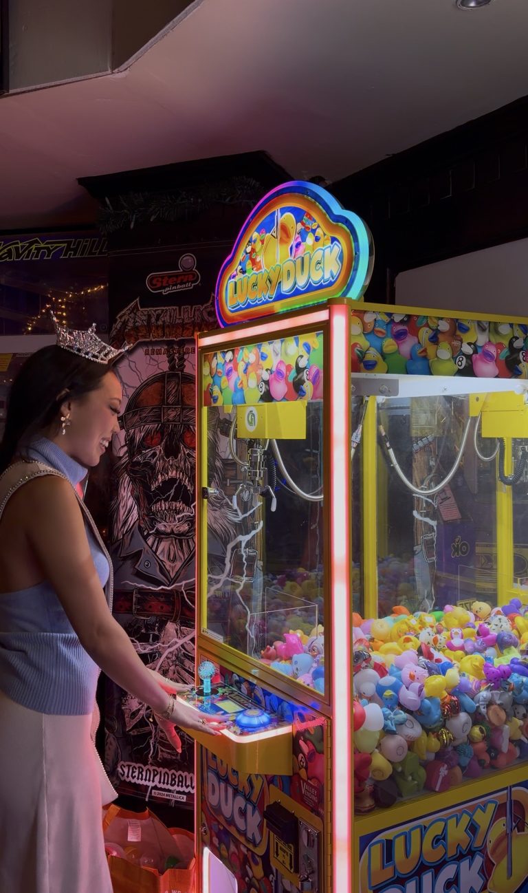A woman plays a brightly lit Lucky Duck claw machine filled with toy ducks, surrounded by arcade decorations and pinball machines, as Miss New York explores Albany NY by Amtrak.