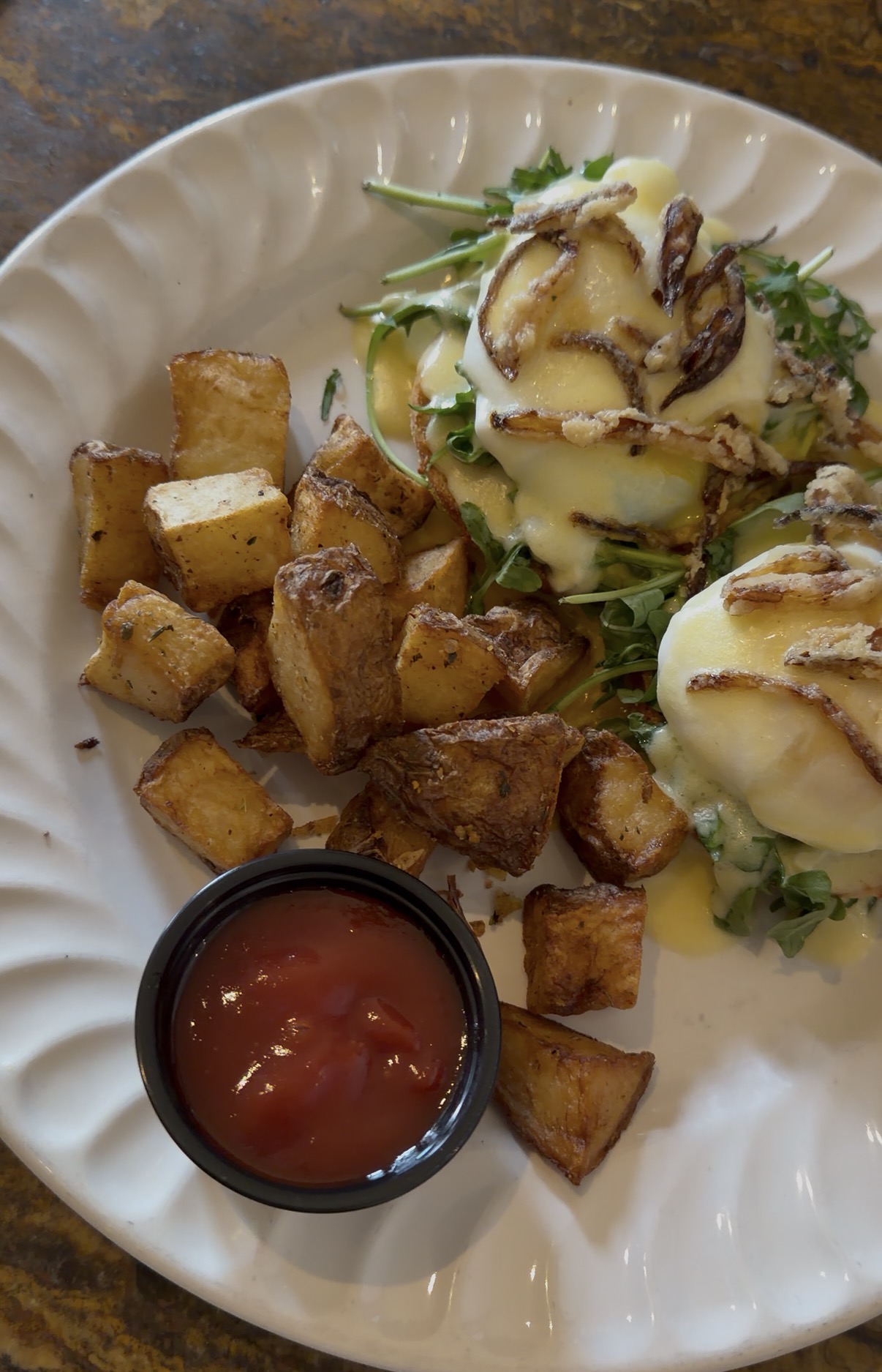 A plate with roasted potato cubes, a side of ketchup, and two eggs Benedict on greens, topped with hollandaise sauce and caramelized onions—just what Miss New York Explores Albany NY by Amtrak might enjoy.