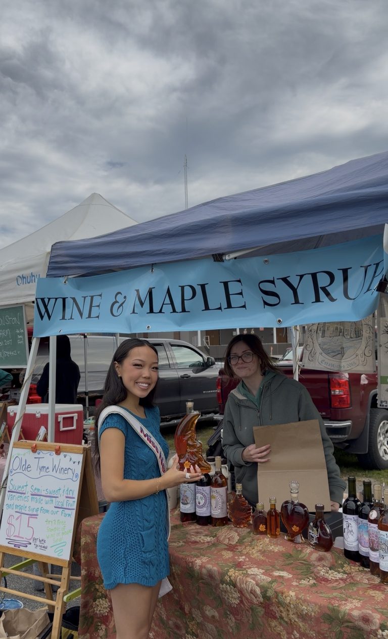 Two people stand at a market booth labeled Wine & Maple Syrup. Miss New York Explores Albany NY by Amtrak, smiling with a maple leaf bottle, while the other stands behind a table of bottles and an Olde Tyme Winery sign.