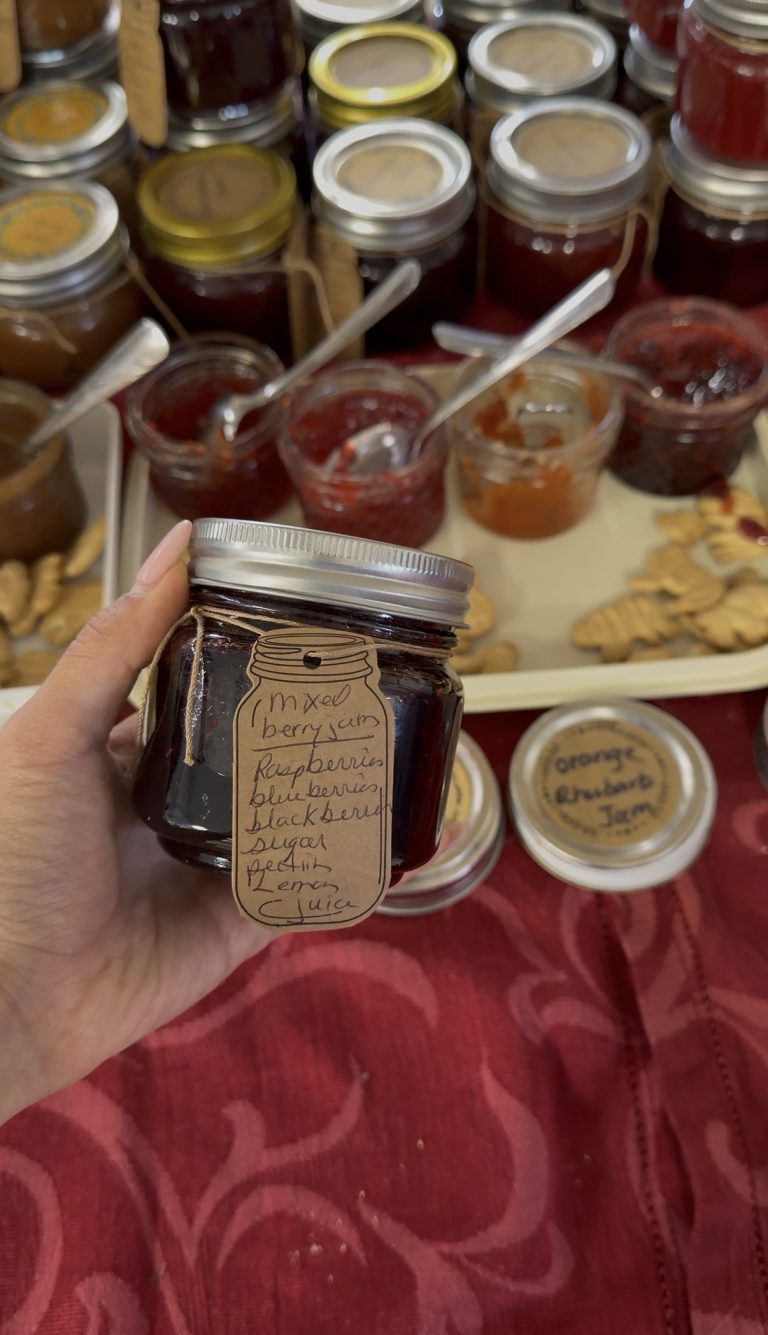A hand holds a jar labeled Mixed Berry Jam with a handwritten tag as Miss New York Explores Albany NY by Amtrak. Other jam jars, sample jars, and crackers sit on a red tablecloth in the background.
