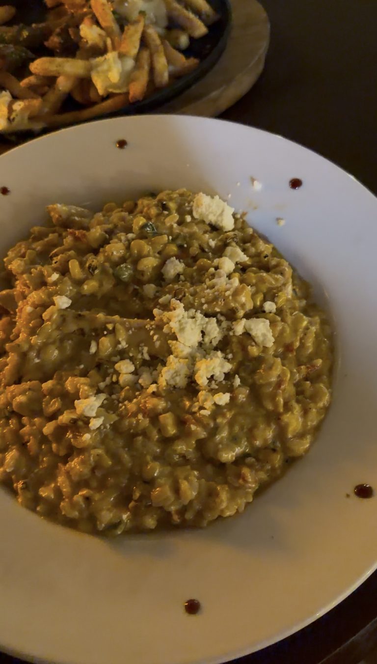A white plate of creamy corn risotto topped with crumbled cheese, with a few sauce dots around the rim—just what Miss New York Explores Albany NY by Amtrak might savor. Fries are visible in the background.