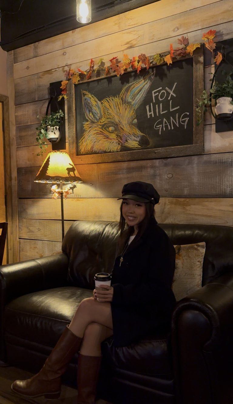 A woman sits on a dark leather couch holding a cup, wearing black and a hat. Behind her, a lamp glows, and a chalkboard reads FOX HILL GANG—Miss New York Explores Albany NY by Amtrak ambiance.