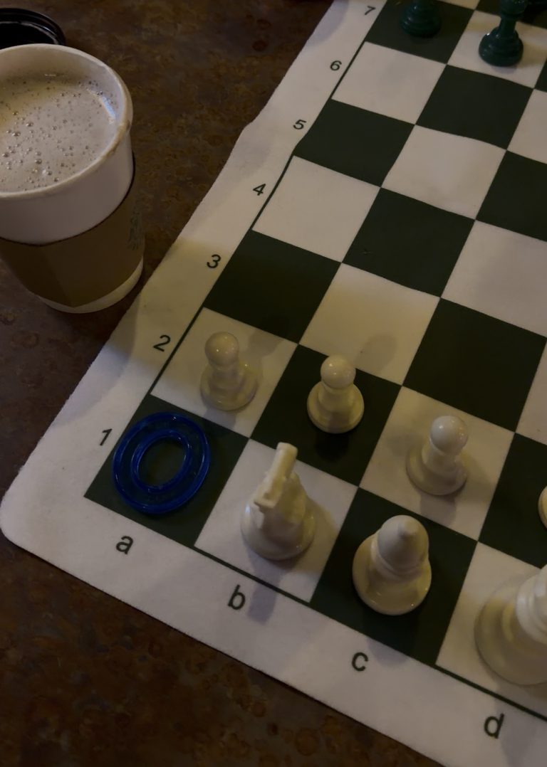 A chessboard with white pieces, a blue ring on a1, and a paper cup of coffee on the left side, all on a brown tabletop—like Miss New York Explores Albany NY by Amtrak, it’s poised for adventure.