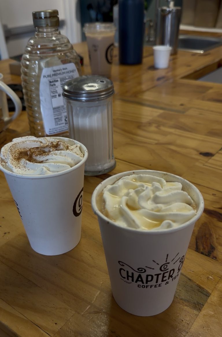 Two cups of coffee with whipped cream sit on a wooden table near a sugar dispenser and honey bottle, as Miss New York Explores Albany NY by Amtrak in the cozy café background.