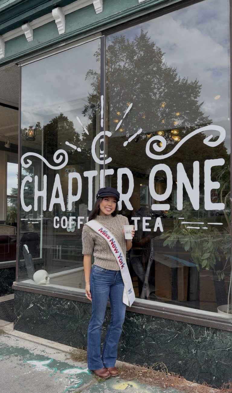 A woman wearing a Miss New York sash stands smiling in front of a window reading Chapter One Coffee & Tea, as Miss New York explores Albany NY by Amtrak. She’s casually dressed and wearing a hat.