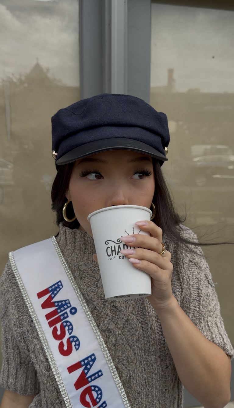 Woman wearing a navy hat, knit sweater, and MISS sash sips from a white to-go coffee cup, looking to the side while outdoors—Miss New York explores Albany NY by Amtrak.