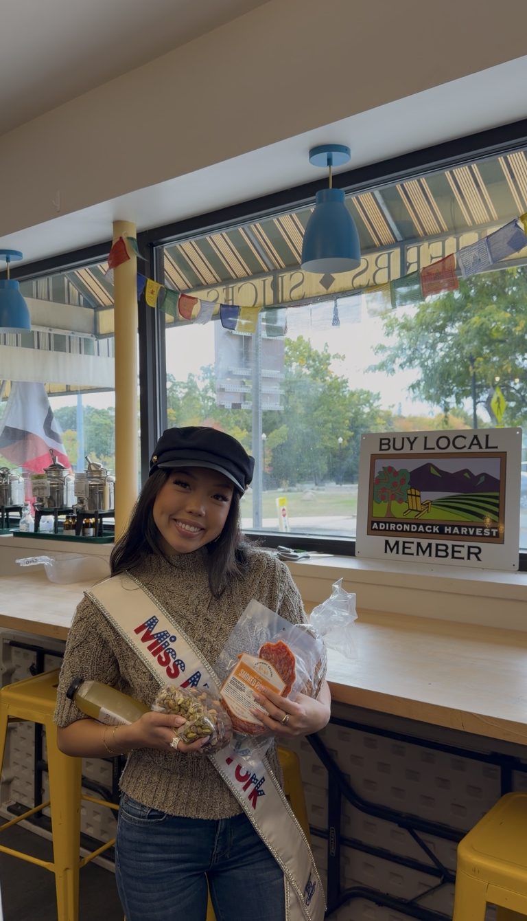 A smiling woman wearing a Miss sash and black cap holds bread and snacks in a café with a Buy Local Adirondack Harvest Member sign, as Miss New York Explores Albany NY by Amtrak.