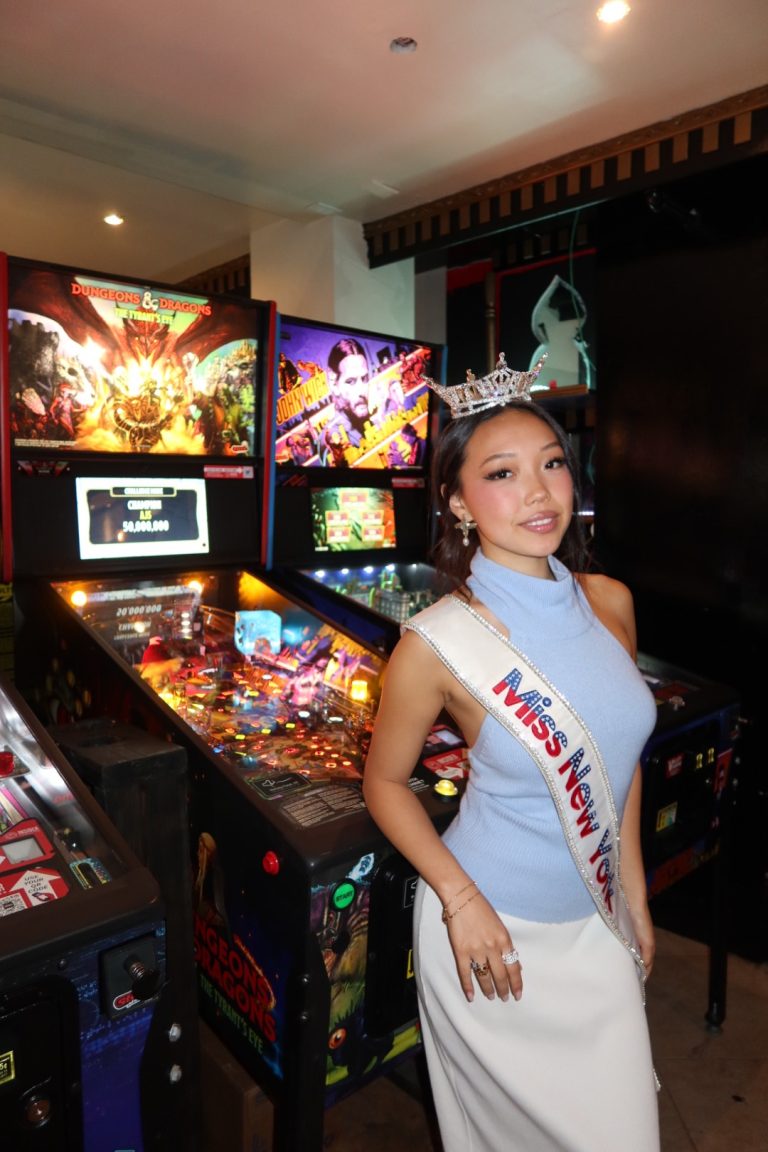 A woman wearing a Miss New York sash and crown poses in front of arcade pinball machines, smiling at the camera as Miss New York explores Albany NY by Amtrak.