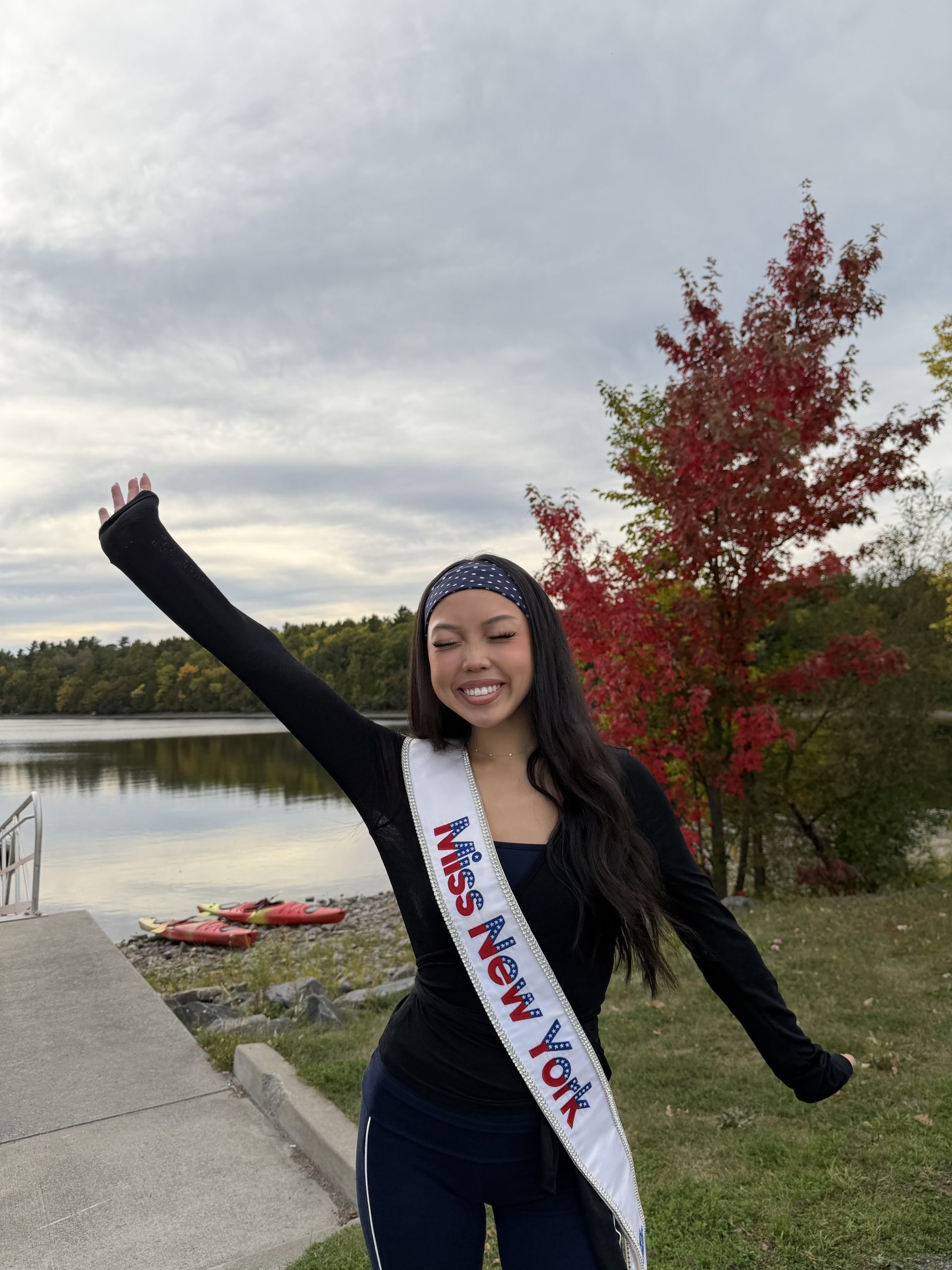 A young woman wearing a Miss New York sash smiles with one arm raised, standing by a lake with colorful autumn trees as Miss New York explores Albany NY by Amtrak.