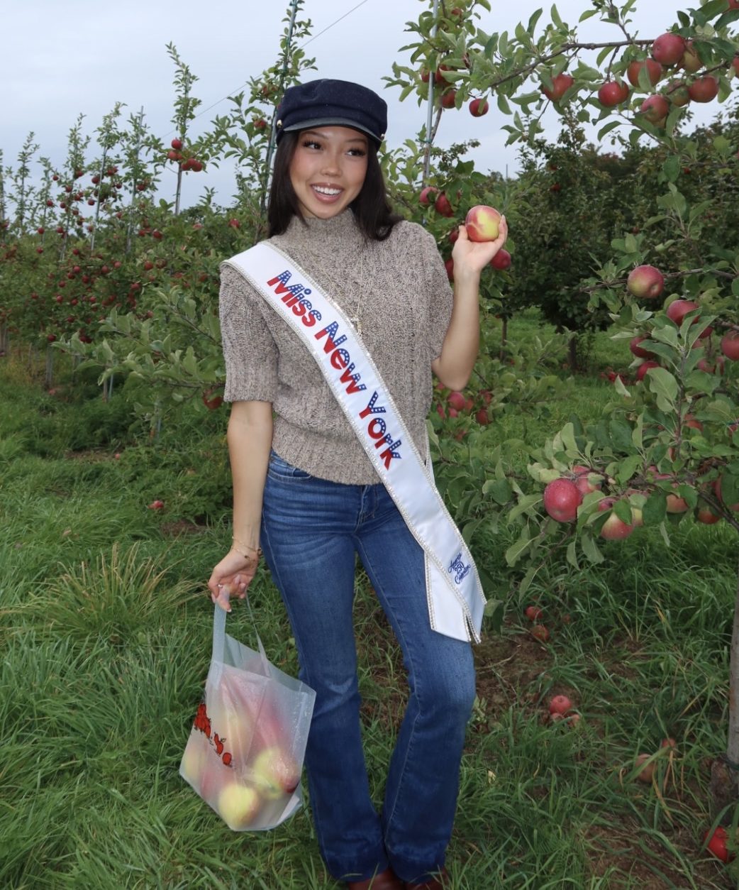 A woman wearing a Miss New York sash, a blue cap, and jeans smiles while holding apples in an orchard as Miss New York explores Albany NY by Amtrak.