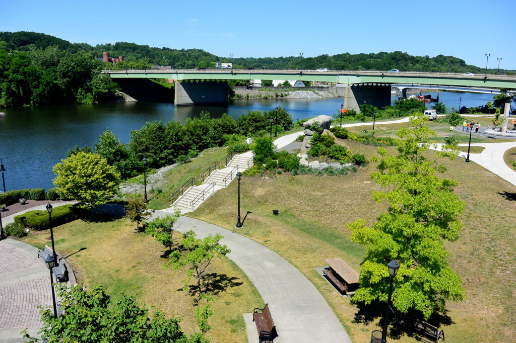Riverlink Park is a riverside park with winding paths, green trees, benches, and a stone staircase. A bridge crosses the river in the background under a clear blue sky.