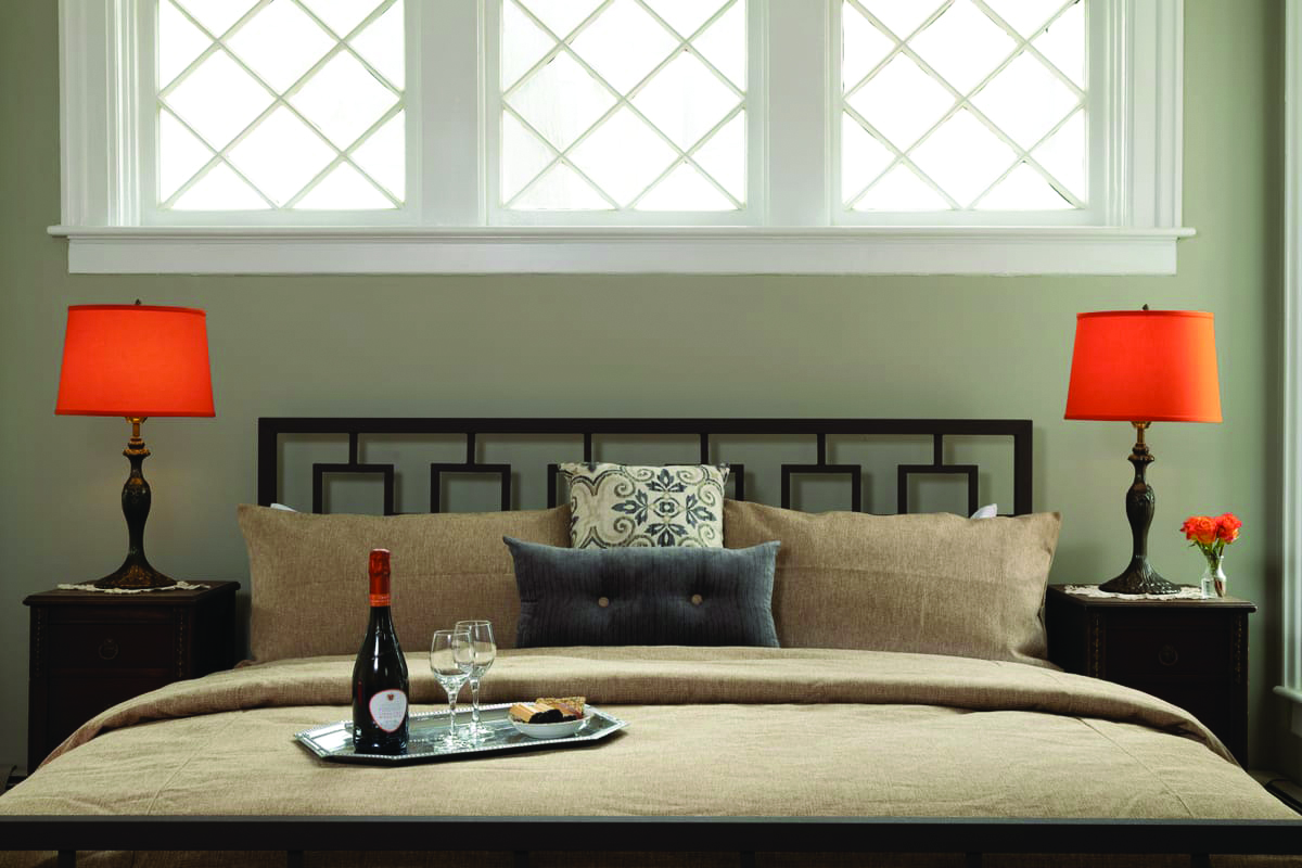 A neatly made bed with tan bedding sits beneath three windows with white frames at the Rosemont Inn Bed & Breakfast. Two orange lamps rest on bedside tables, while a tray with wine and glasses and three decorative pillows complete the inviting scene.