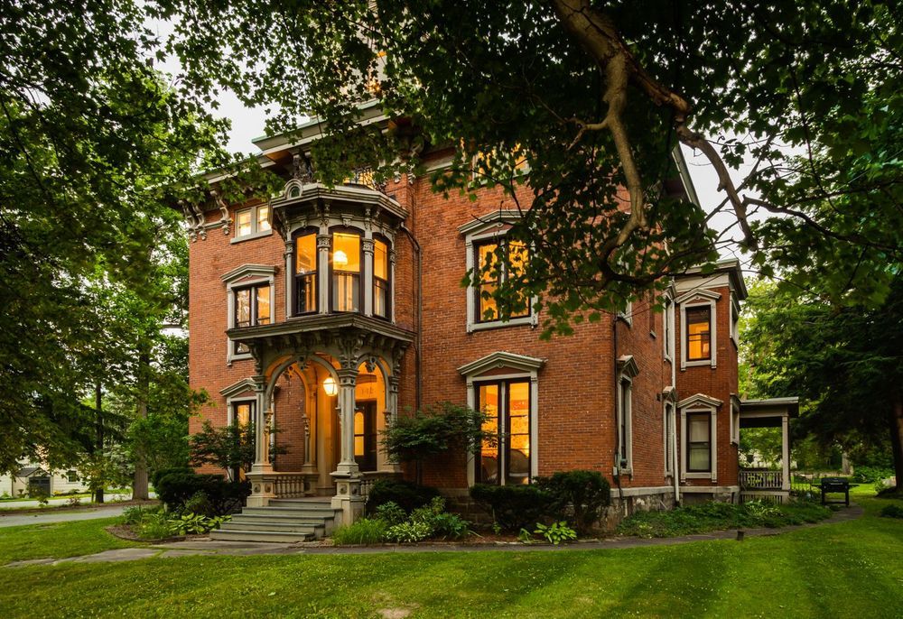 A large, historic red-brick house with ornate details and glowing windows, reminiscent of the charm found near Chelsea Market, stands surrounded by lush green trees and a well-kept lawn at dusk.