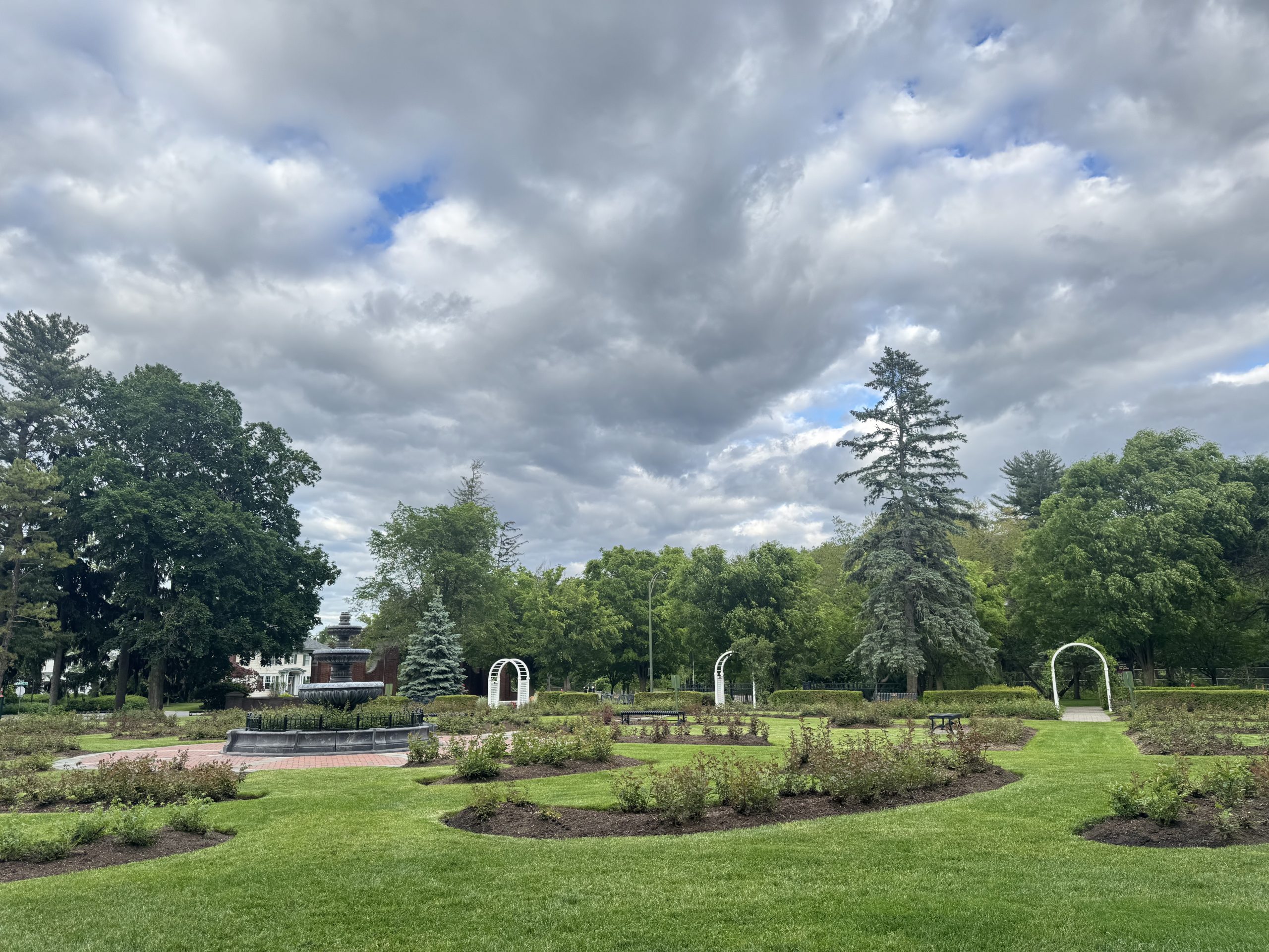 A landscaped garden with circular flowerbeds, white archways, a central fountain, and tall trees under a cloudy sky evokes the elegance of the Schenectady Rose Garden.