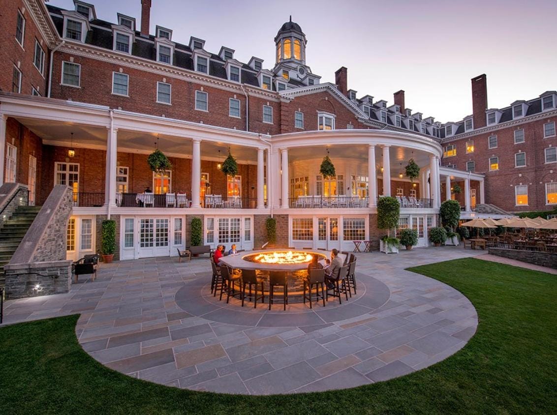 A grand brick hotel with large windows and columns overlooks a spacious patio, reminiscent of the stately charm found near the Hudson-Athens Lighthouse. A group of people sit around a circular outdoor fire pit as dusk falls, with trimmed lawns and elegant outdoor seating nearby.