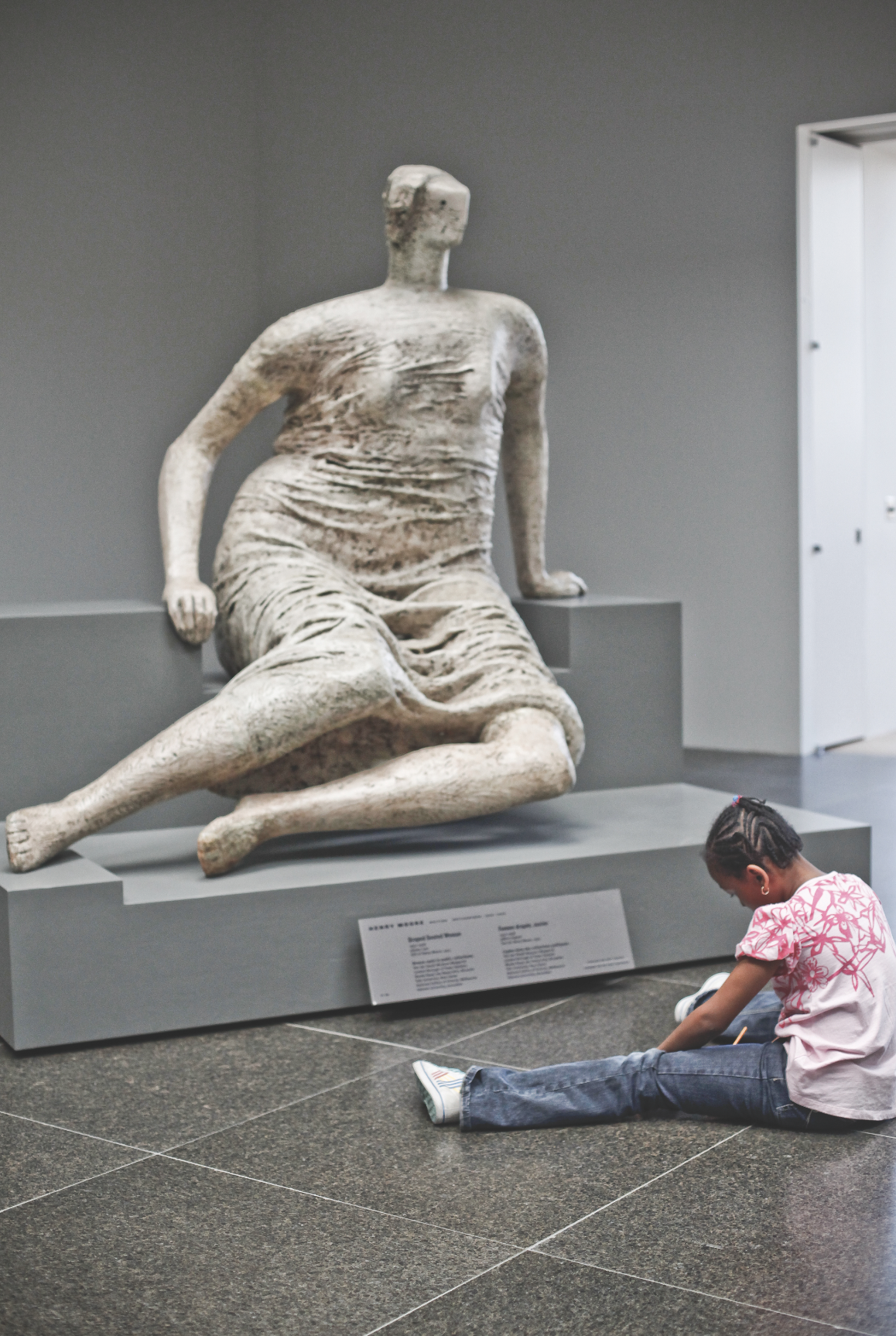 A young girl sits on the floor in front of a large, abstract, seated sculpture at the Art Gallery of Ontario, mimicking the statue’s pose.