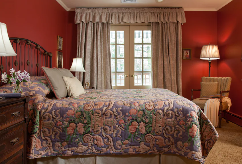 A cozy bedroom at Journey Inn Bed & Breakfast features red walls, a bed covered by a floral quilt, bedside tables with lamps, an armchair, and large windows with beige curtains letting in daylight.