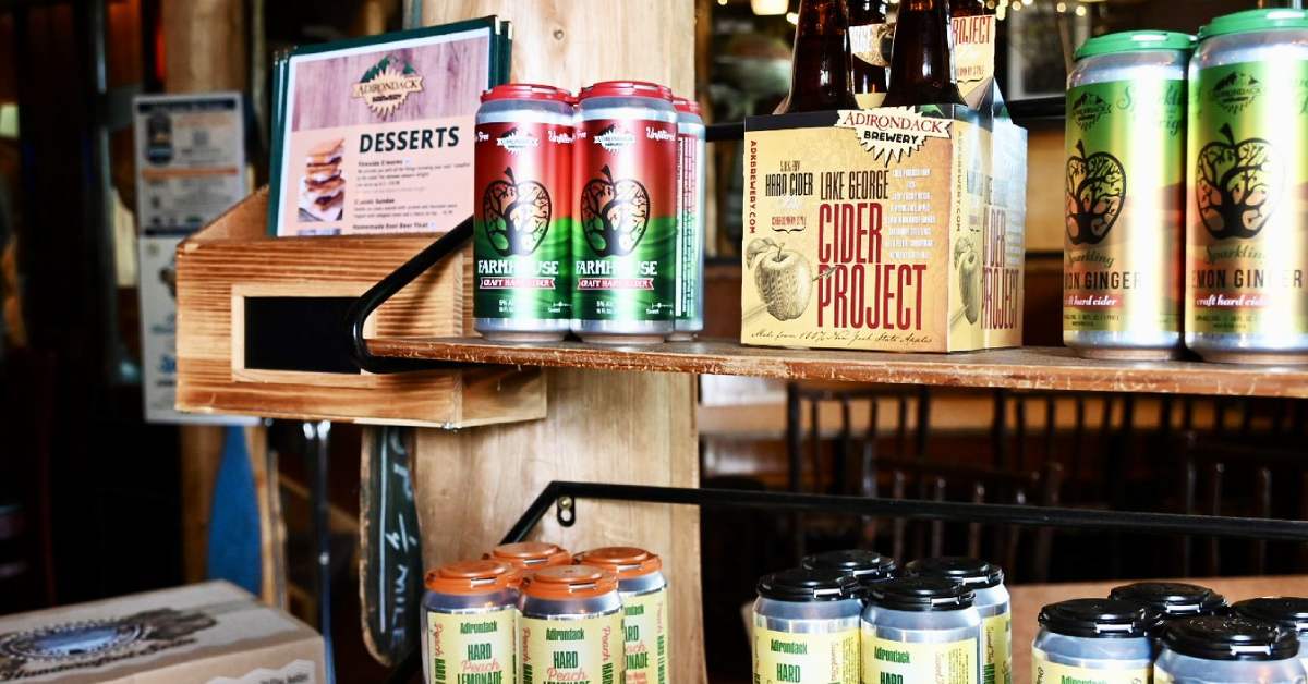 Shelves display assorted craft beer cans, a boxed cider project beer, and a dessert menu in a cozy, rustic bar setting with wood accents and soft lighting—perfect for those exploring the Adirondack Craft Beverage Trail.