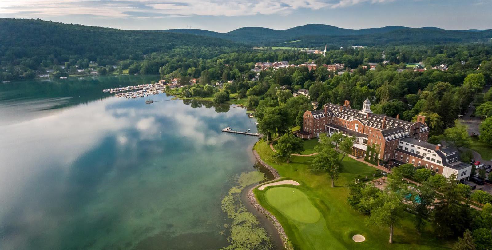 Aerial view of The Otesaga Resort Hotel, a large lakeside hotel surrounded by trees, with a golf course, docks, and a small town in the background near forested hills.