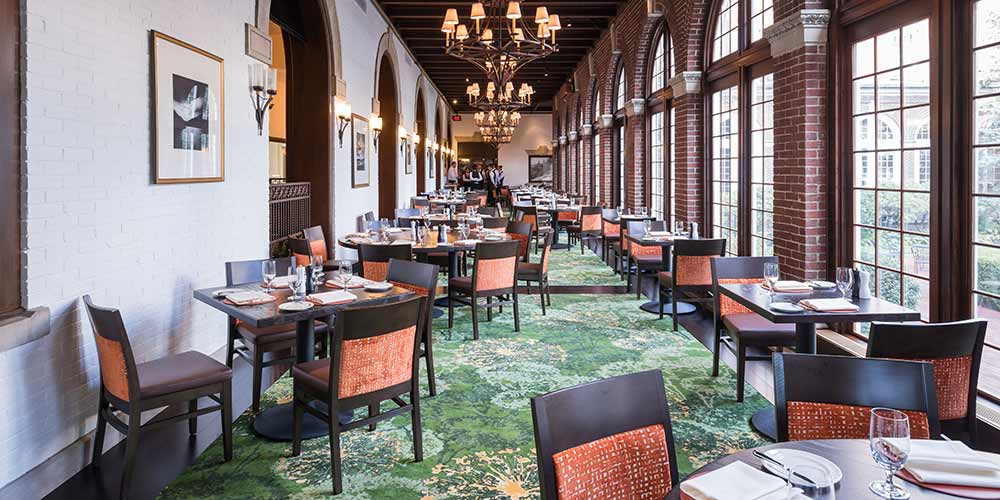 Elegant restaurant with arched windows, brick walls, green patterned carpet, and neatly set tables with black chairs. Chandeliers hang from a wooden beamed ceiling, and sunlight fills the bright, spacious dining area—an inviting ambiance that defines American Bounty Restaurant.