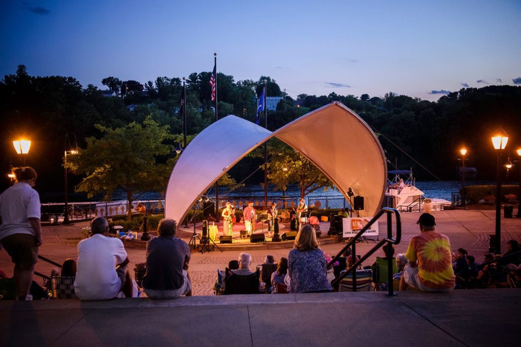 People sit on outdoor steps at dusk in Riverlink Park, watching a band perform under a white canopy by the river. Streetlights glow, and trees line the background.