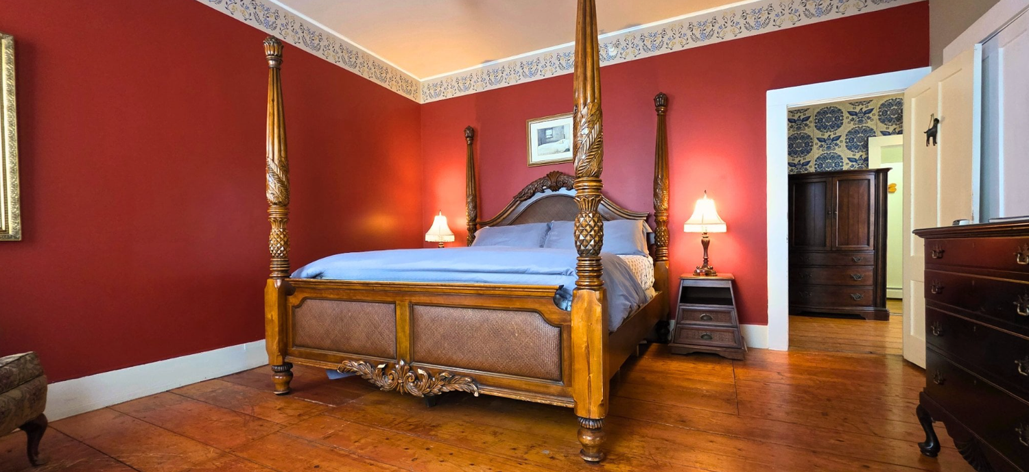A bedroom at the Rosemont Inn Bed & Breakfast features striking red walls, a wooden four-poster bed, two bedside tables with lamps, polished wood floors, and an open door revealing another room adorned with elegant blue wallpaper.