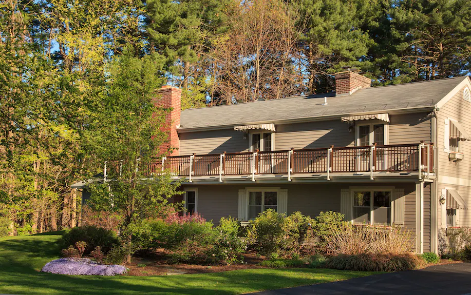 Two-story gray house with white trim, large balcony, and awnings—The Paw House Inn is surrounded by green lawn, flowering shrubs, and trees, with sunlight filtering through.