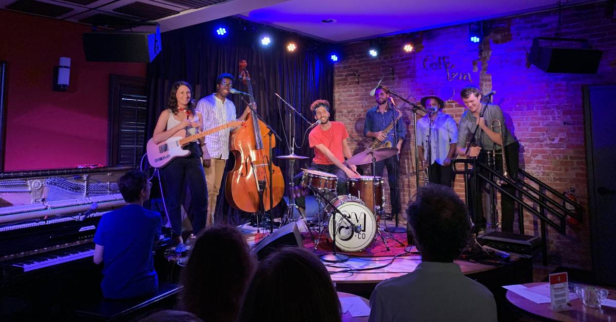 A jazz band performs on stage with piano, guitar, double bass, drums, saxophone, trombone, and trumpet, as an audience watches in a cozy, brick-walled venue with purple lighting reminiscent of the enchanting evenings at the Schenectady Rose Garden.