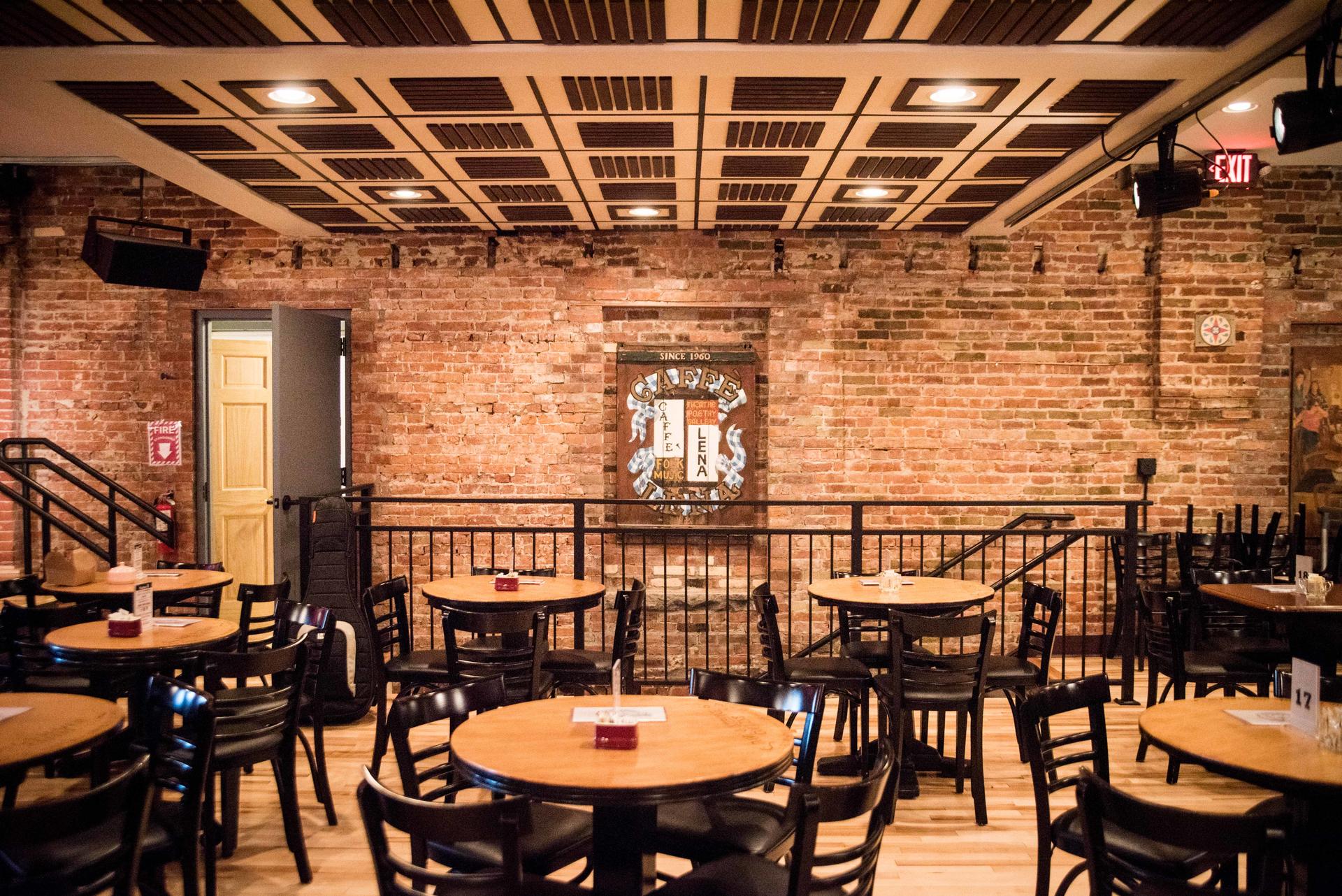 A cozy comedy club with brick walls, round wooden tables, black chairs, and a small stage area under warm ceiling lights—just a short stroll from the vibrant beauty of the Schenectady Rose Garden.