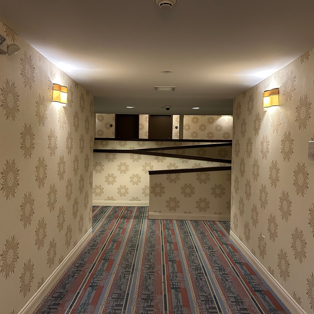 A hallway with patterned wallpaper and striped carpet ends abruptly at a half-wall, leading to an unusual, seemingly inaccessible area with cabinets above—reminiscent of the quirky interiors found at Hotel Skyler Syracuse. Two wall lights illuminate the corridor.